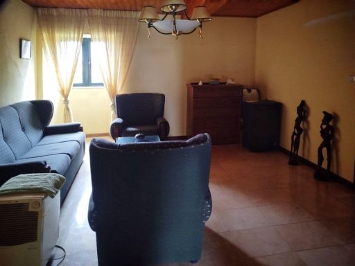 Salón con sofá y sillón, ventana con cortinas, y muebles de madera en tonos cálidos.
